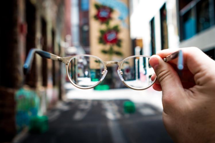 A person holds up a pair of glasses in front of a street that is out of focus
