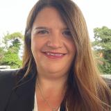 Photograph of a white woman who has long light brown hair, dark brown eyes set to a background of green trees.